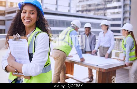 Alles ist bis zum Code. Zugeschnittenes Porträt einer attraktiven jungen Bauarbeiterin, die draußen mit ihrem Team im Hintergrund steht. Stockfoto