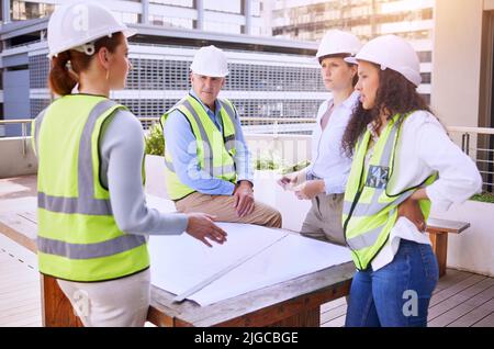 Kommunikation ist wichtig in der Bauindustrie. Eine Gruppe von Bauarbeitern schaut auf Baupläne, während sie draußen auf der Baustelle stehen. Stockfoto