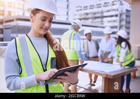 Die Bauarbeiten laufen. Eine attraktive junge Bauarbeiterin, die ein Tablet benutzt, während sie draußen mit ihrem Team im Hintergrund steht. Stockfoto