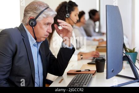 Ich könnte diesen Verkauf einfach verlieren. Ein reifer Callcenter-Agent, der gestresst aussagt, während er in einem Büro mit seinen Kollegen im Hintergrund arbeitet. Stockfoto