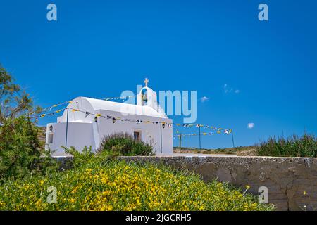 Weiße griechisch-orthodoxe Kapelle oder Kirche auf dem Hügel gegen klaren blauen Himmel an sonnigen Tagen. Bunte Blumen und Sträucher im Vordergrund. Milos, Griechenland Stockfoto