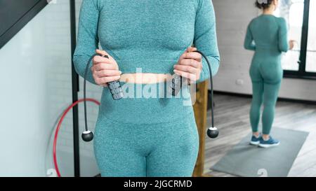 Home Fitness Workout Konzept. Kabelloses Springseil in den Händen einer Frau in Sportkleidung auf dem Hintergrund einer Ziegelwand. Moderne Technologien im Sport Stockfoto