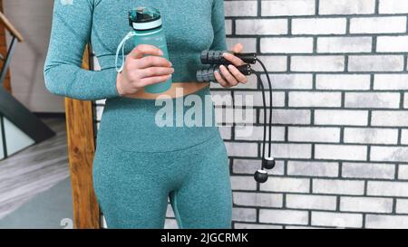 Frau in Sportkleidung hält in ihren Händen ein kabelloses Springseil und eine Wasserflasche. Home Fitness Workout Konzept. Moderne Technologien im Sportbereich Stockfoto