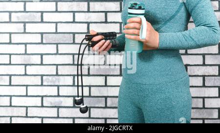 Kabelloses Springseil und eine Wasserflasche in den Händen einer Frau in Sportkleidung auf dem Hintergrund einer Ziegelwand. Home Fitness Workout Konzept. Sport AC Stockfoto