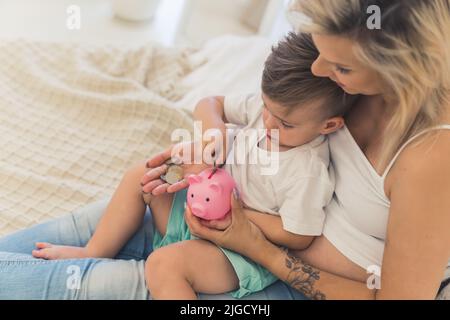 Die junge, tausendjährige blonde kaukasische Mutter in einer weißen Beschnittplatte hält ihren kleinen Sohn auf den Schoß und lehrt ihn, wie man mit dem Sparschwein Geld spart. Hochwertige Fotos Stockfoto