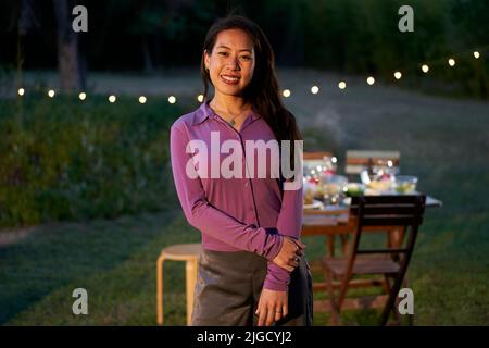 Porträt einer asiatischen Kellnerin, die die Kamera anschaut. Die chinesische Restaurantarbeiterin steht mit einem netten Lächeln auf der Terrasse vor einem Tisch Stockfoto