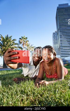 Vertical eines fröhlichen jungen afroamerikanischen Paares, das ein Selfie-Portrait mit seinem Smartphone auf dem Rasen im Stadtpark aufnahm. Lächelnder Junge Stockfoto
