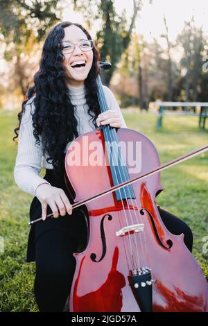 Glückliche junge Brünette Frau mit Brille lachen und spielen Cello bei Sonnenuntergang im Park. Vertikal Stockfoto