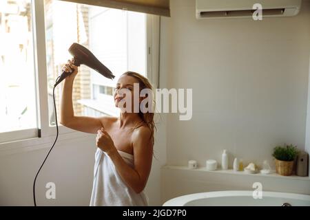 Glücklich fröhlich junge hübsche Frau in Handtuch trocknet Haar mit Trockner nach dem Baden. Stockfoto