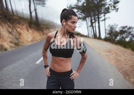 Selbstbewusste Sportlerin, die in Sportkleidung auf der Straße steht und wegschaut. Fit junge Frau mit einem Outdoor-Training. Stockfoto