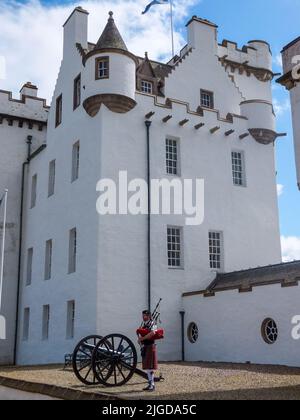 Das mittelalterliche Blair Castle steht in Weiß über einem antiken Kanon und einem Dudelsackspieler in Pitlochry, Perthshire, Schottland, Großbritannien. Stockfoto