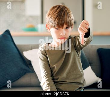 Kein Kind genießt es, allein und hungrig zu sein. Porträt eines niedlichen kleinen Jungen, der die Daumen nach unten zeigt, während er zu Hause auf einem Sofa sitzt. Stockfoto