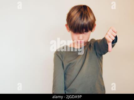 Er muss in seinen Sinn für Humor hineinwachsen. Studioaufnahme eines niedlichen kleinen Jungen, der die Daumen gegen eine Wand zeigt. Stockfoto