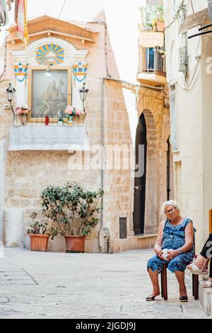 Ältere lokale Frau sitzt auf einem Stuhl vor der Haustür in einer engen Straße der Altstadt von Bari, Apulien, Italien, vertikal Stockfoto