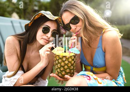Freundinnen trinken Saft direkt aus einer geschnittenen Ananas mit Stroh Blick auf die Kamera - Sommer Lifestyle Menschen Konzept - zwei junge Frau trinken Stockfoto