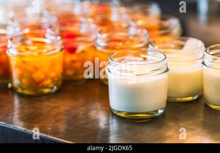 Hausgemachter Joghurt in kleinem Flaschenglas auf Edelstahlregal mit verschwommenem Hintergrund von Obstsalat in kleinem Flaschenglas, Nahaufnahme mit Stockfoto