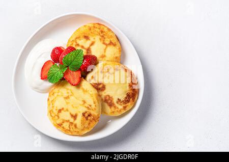 Hüttenkäse-Pfannkuchen mit saurer Rahm und Beeren auf grauem Hintergrund. Stockfoto