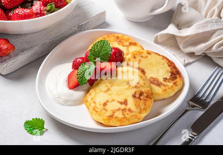Quark-Pfannkuchen mit saurer Creme und Erdbeeren auf hellem Hintergrund. Stockfoto