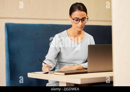 Shes im Geschäft des Names. Eine attraktive reife Geschäftsfrau, die Notizen macht, während sie an einem Schreibtisch im Büro an ihrem Laptop arbeitet. Stockfoto