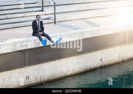 Ein Geschäftsmann, der in Tauchausrüstung am Rande eines Wasserkörpers sitzt. Stockfoto