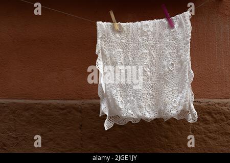 Weiße gehäkelte Tischdecke, zum Aufhängen am Draht aufgehängt, zum Trocknen nach dem Waschen. Hintergrund mit brauner Wand. Stockfoto