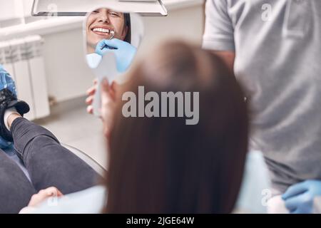 Glückliche Patientin schaut im Spiegel auf ihre Zähne und lächelt Stockfoto