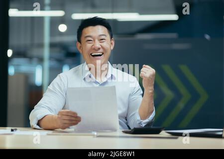 Porträt eines erfolgreichen asiatischen Geschäftsmannes, lächelnder Mann und Blick auf die Kamera, Brief und Dokument in den Händen haltend, glücklich mit Erfolg, die Hand nach oben Geste des Triumphes und des Sieges haltend Stockfoto