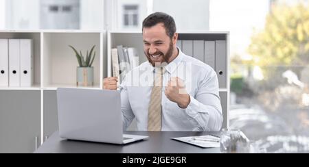 Das Rampenlicht leuchtet hell auf seinen Erfolg. Ein junger Geschäftsmann jubelt, während er in einem Büro an einem Laptop arbeitet. Stockfoto