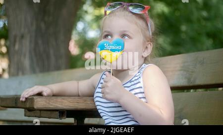 Das kleine blonde Mädchen hält Lebkuchen in ukrainischen Nationalfarben in der Hand und sagt: „Die Ukraine ist ich“. Nahaufnahme Porträt des Mädchens auf Park benc sitzen Stockfoto