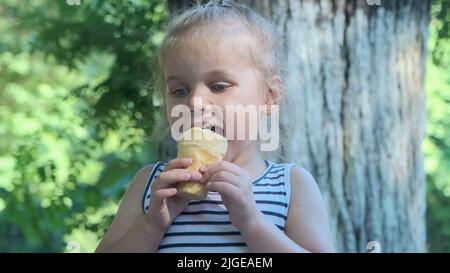 Nettes kleines Mädchen isst draußen Eis. Nahaufnahme eines blonden Mädchens, das auf der Parkbank sitzt und Eis isst. Stockfoto