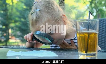 Kleines Mädchen schaut vorsichtig in die Linse auf das Salz. Nahaufnahme des blonden Mädchens studiert Salzkristalle, während sie durch die Lupe betrachtet wird Stockfoto