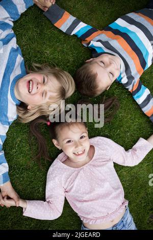 Glücklich zwei Brüder und Schwester liegen auf dem Gras mit einem Gefühl von Glück und Einheit. Gute Laune, unbeschwerte, fröhliche Kindheit. Blick von oben Stockfoto