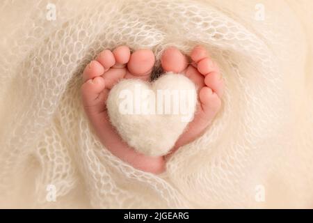 Gestricktes weißes Herz in die Beine eines Babys. Weiche Füße in einer weißen Decke. Nahaufnahme von Zehen, Fersen und Füßen eines Neugeborenen. Stockfoto