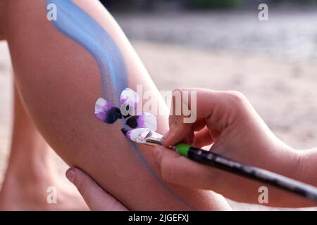 Künstler malt eine Blume im Bein einer Frau am Strand Stockfoto