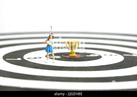 Miniatur Menschen Spielzeug Figur Fotografie. Ein Golfermädchen, das über der Dartscheibe mit Goldtrophäe in der Mitte steht, isoliert auf weißem Hintergrund. Bild-ph Stockfoto