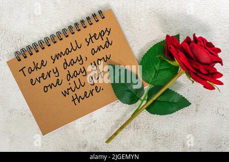 Motivationszitat auf braunem Notizbuch mit roten Rosen auf Marmorschreibtisch - machen Sie jeden Tag kleine Schritte, und eines Tages kommen Sie dorthin. Stockfoto
