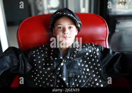 Porträt eines Kindes in Lederjacke und Mütze auf einem Stuhl sitzend Stockfoto