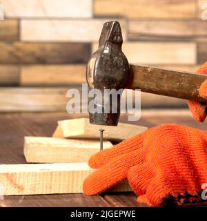 Nahaufnahme eines Zimmermanns in orangefarbenen Arbeitshandschuhen hämmert einen Nagel in ein Holzbrett. Stockfoto