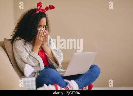 Allein verbrachte Weihnachtsfeiertage schlagen hart zu. Eine junge Frau, die zu Hause einen Laptop benutzt und während der Weihnachtszeit auf dem Sofa weint. Stockfoto