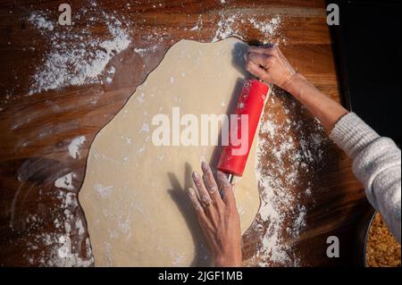 Ansicht von oben, wie weibliche Hände mit einem Nudelholz auf dem heimischen Esstisch hausgemachten Teig Rollen. Stockfoto