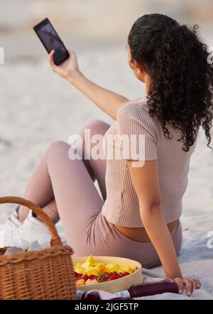 Einige meiner Lieblingsmomente am Strand festhalten. Eine unkenntliche Frau, die allein am Strand sitzt und mit ihrem Handy Selfies macht. Stockfoto