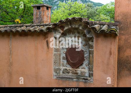 Eine runde Metallplatte ist in eine Wandnische eines Hauses in Mourèze, Hérault, Frankreich, eingelassen, geschützt von einem kleinen Ziegeldach. Der Zweck der Platte ist nicht identifizierbar. Stockfoto