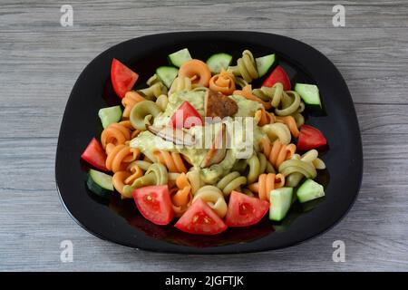 Tricolore Pasta mit Steinpilzen, frischem Gemüse und saurer Sauce serviert in schwarzer Keramikplatte auf grauem Holzhintergrund Stockfoto