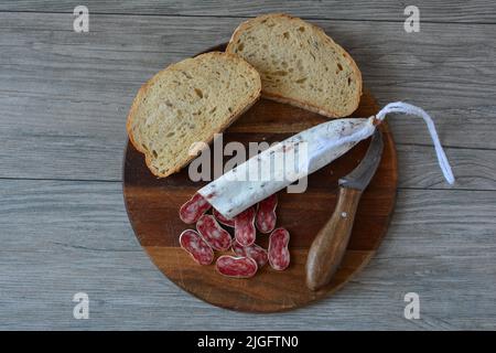 Sehr würzige spanische Schweinewurst mit weißem Schimmel, auf dunklem Holzschneidebrett geschnitten, mit etwas dunklem Brot und altem rustikalem Messer, Draufsicht Stockfoto