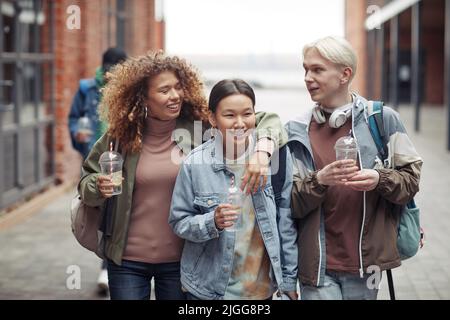 Drei junge, fröhliche Freunde in Casualwear trinken in Plastikgläsern und diskutieren lustige Dinge auf dem Weg nach Hause nach dem College Stockfoto