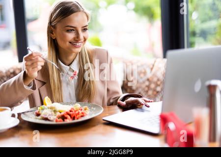 Nachdenkliche junge Geschäftsfrau, die auf der Freilichtterrasse des Dachrestaurants zu Mittag gegessen hat und auf den Bildschirm ihres Laptops blickt Stockfoto