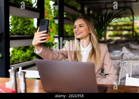 Schöne Geschäftsfrau, die mit einem Smartphone Selfie gemacht und an ihrem Laptop gearbeitet hat, während sie tagsüber in einem Café sitzt Stockfoto