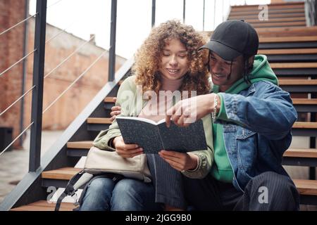 Zwei Jugendliche Studenten sitzen auf der Treppe und lesen Buch oder durch Vortragsnotizen, während glücklicher Kerl umarmt seine Freundin Stockfoto