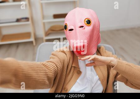 Lustige blonde Dame in rosa Fischmaske warmen Pullover tun Selfie verwenden Telefon sitzen im Sessel im modernen Zuhause Interieur. Pause von der Arbeit, Pause machen Stockfoto