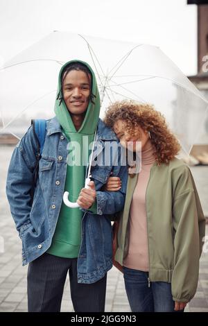 Glückliches junges interrassisches Paar in Casualwear, das unter einem Regenschirm dicht beieinander steht und beim Gehen auf die Kamera schaut Stockfoto